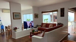 A living room with a sofa, coffee table, and TV. There is a window with a view of the outside. - 4 suites, air conditioning, TV, internet, swimming pool, 150 meters from the beach! Pets allowed! (Guarujá)