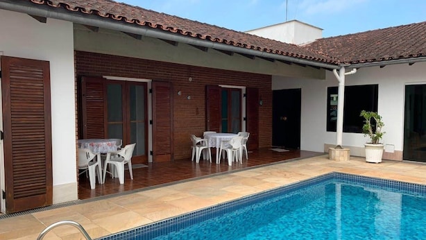 A swimming pool with a tiled deck, a covered patio area with tables and chairs, and a brick building with wooden shutters. - 4 suites, air conditioning, TV, internet, swimming pool, 150 meters from the beach! Pets allowed! (Guarujá)
