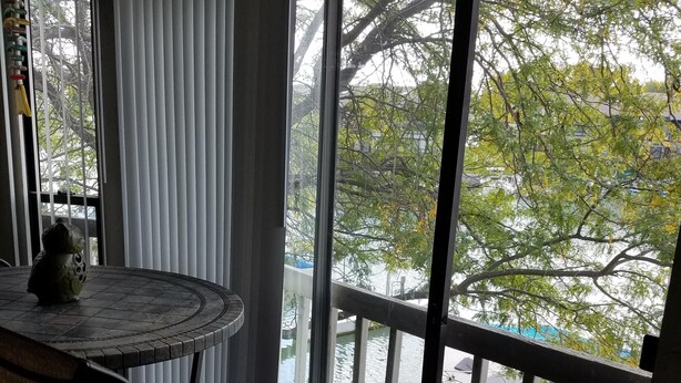 A room with a round table and chairs, large windows with blinds, and a view of a body of water and trees. - Lake Erie Green Cove Getaway (Oak Harbor)