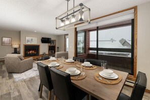 Un coin repas avec une table en bois dressée pour quatre personnes, une cheminée et une grande fenêtre donnant sur un quai.