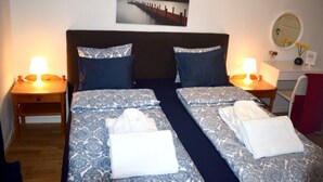 Uma cama de casal com roupa de cama estampada, duas luminárias de cabeceira, um criado-mudo, um espelho e um quadro na parede.