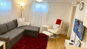 Uma sala de estar moderna com um sofá seccional cinza, um tapete vermelho, uma poltrona branca, uma mesa redonda pequena e uma TV na parede.