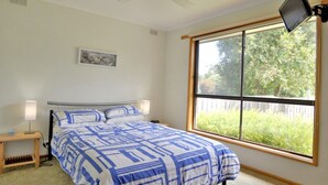 Ein Schlafzimmer mit Bett, Nachttisch, Lampe, Deckenventilator, einem Fenster mit Blick auf Grünflächen und einem an der Wand montierten Fernseher.