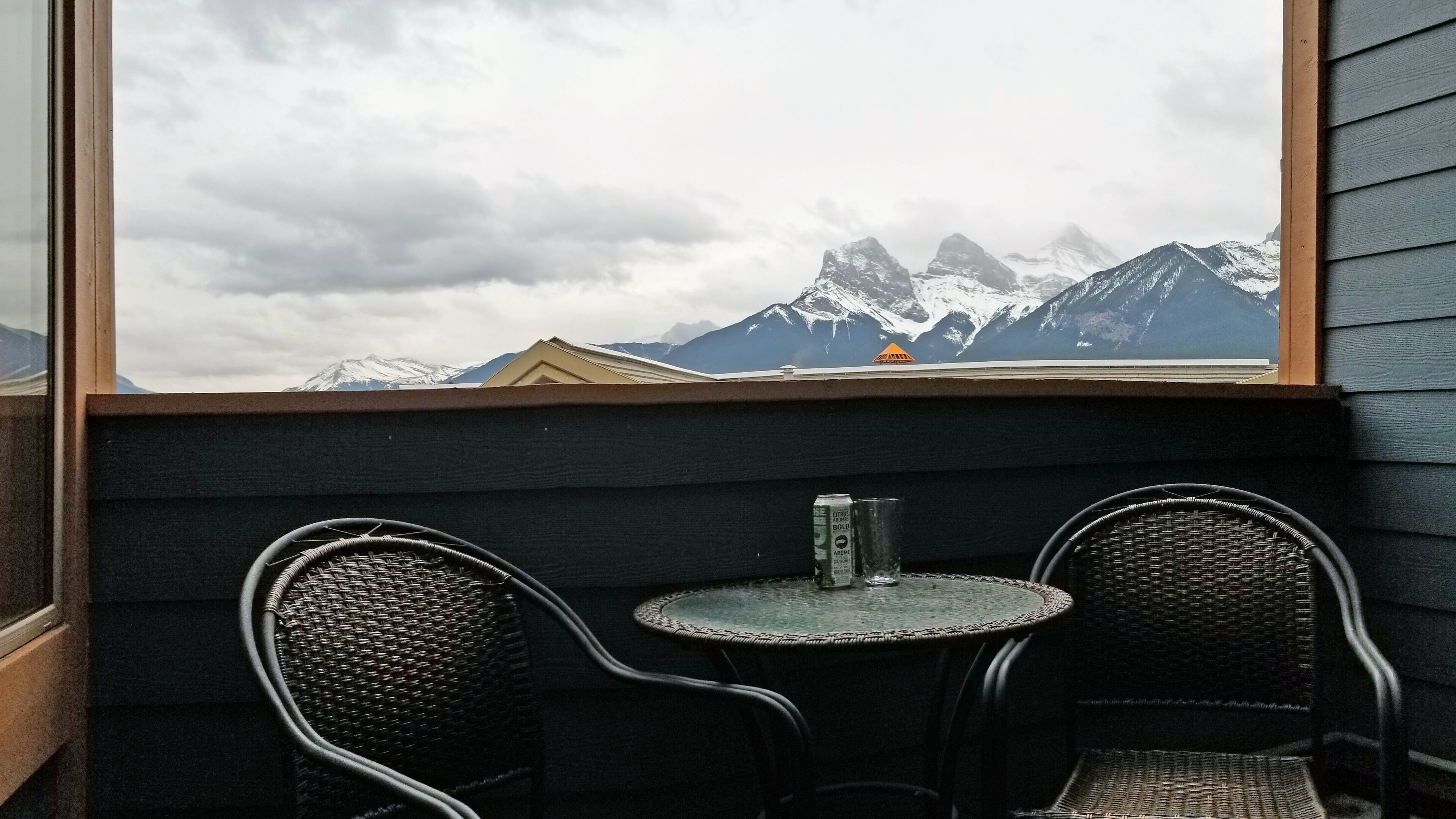 Uma pequena área externa com assentos, incluindo duas cadeiras de vime e uma mesa, com vista para uma cordilheira com picos nevados.