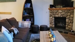 Un salon avec une cheminée en pierre, un canapé marron agrémenté de coussins, une table basse en bois avec des lanternes, et une bibliothèque adossée au mur.