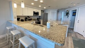 A modern kitchen with granite countertops, white cabinetry, a stainless steel microwave, and a built-in oven. There are three white bar stools and a large island with a sink. - Gulf Front Condo with Stunning Views! (Pensacola)