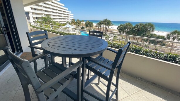 A balcony with a round table and chairs, overlooking a beach and a hotel building. - Gulf Front Condo with Stunning Views! (Pensacola)