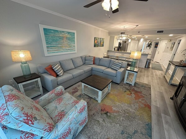 A living room with a sectional sofa, a coffee table, and a patterned armchair. There is a painting on the wall and a ceiling fan. - Gulf Front Condo with Stunning Views! (Pensacola)