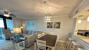 A modern living room with a grey sofa, a wooden dining table with white chairs, and a kitchen area with white cabinets and a granite countertop. - Gulf Front Condo with Stunning Views! (Pensacola)