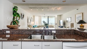 A modern kitchen with granite countertops, stainless steel sink, and a large window offering a view of the living area. - Gulf Front Condo with Stunning Views! (Pensacola)