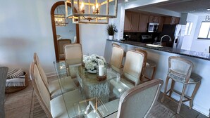 Zona pranzo con tavolo in vetro circondato da sedie in vimini, un lampadario e una cucina sullo sfondo.