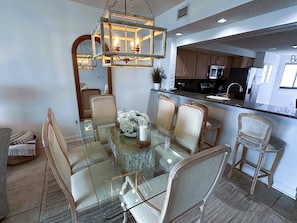 A dining area with a glass table surrounded by wicker chairs, a chandelier, and a kitchen visible in the background.
