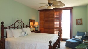 A bedroom with a wooden bed, a ceiling fan, a nightstand with lamps, a window with blinds, and a blue armchair. - Gulf Front Condo with Stunning Views! (Pensacola)