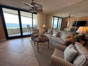 A spacious living room with a large window overlooking the ocean, a ceiling fan, and a television.