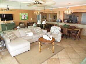 A spacious living room with a sofa, armchairs, a dining table, and a kitchen area visible through a glass partition.