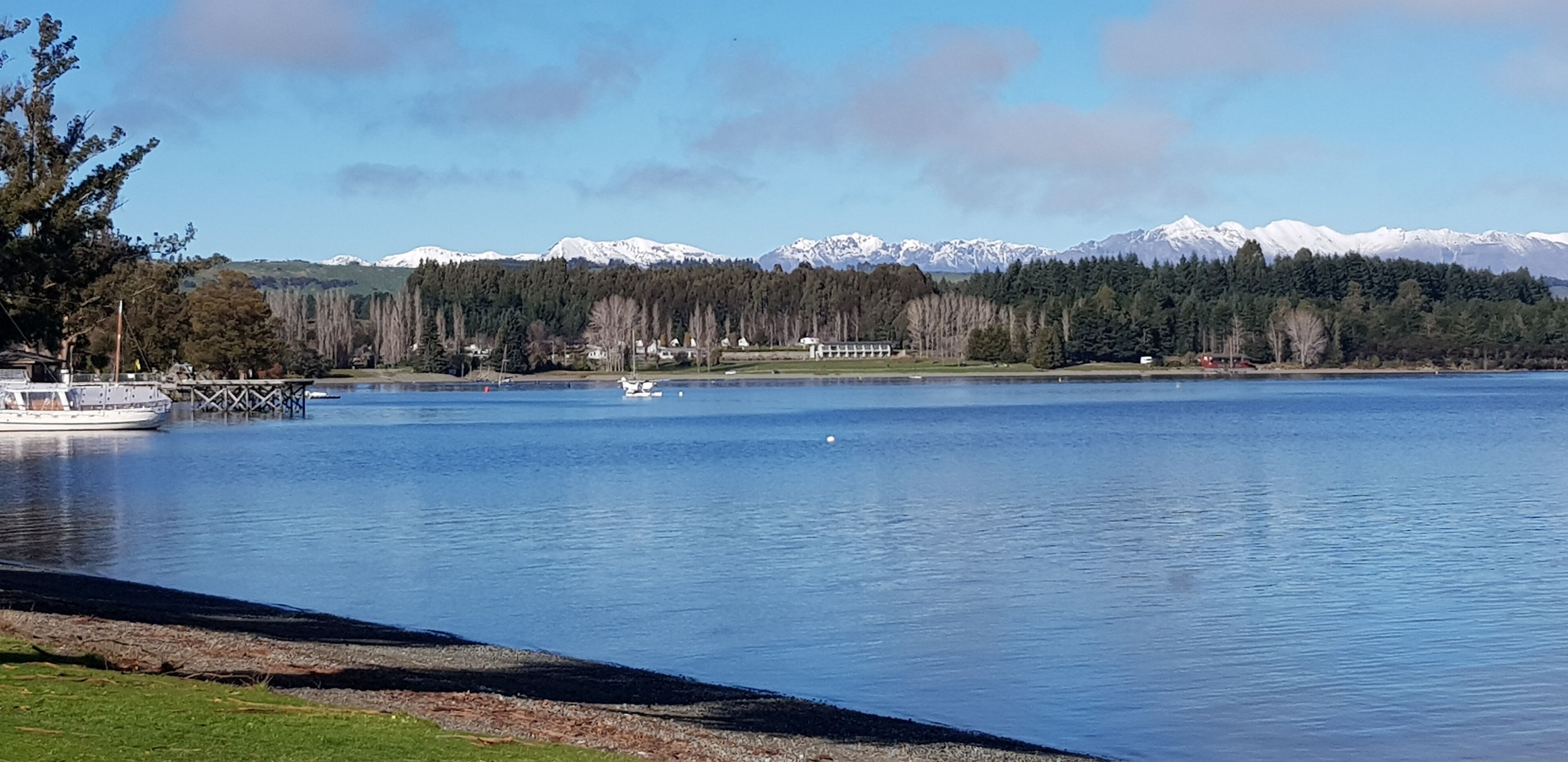 Un lac paisible, avec un quai et un bateau, entouré d’arbres et dominé, au loin, par des montagnes aux sommets enneigés.