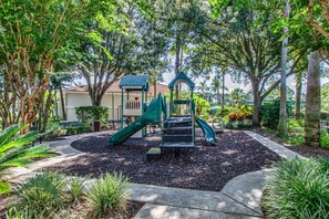 Children can play on the green slide and climbing structure amidst lush trees and shrubs.