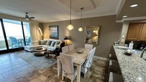 Un vaste espace de vie comprenant un grand canapé, une table à manger et des chaises, une cuisine dotée de plans de travail en granit et un balcon aménagé avec du mobilier d’extérieur.