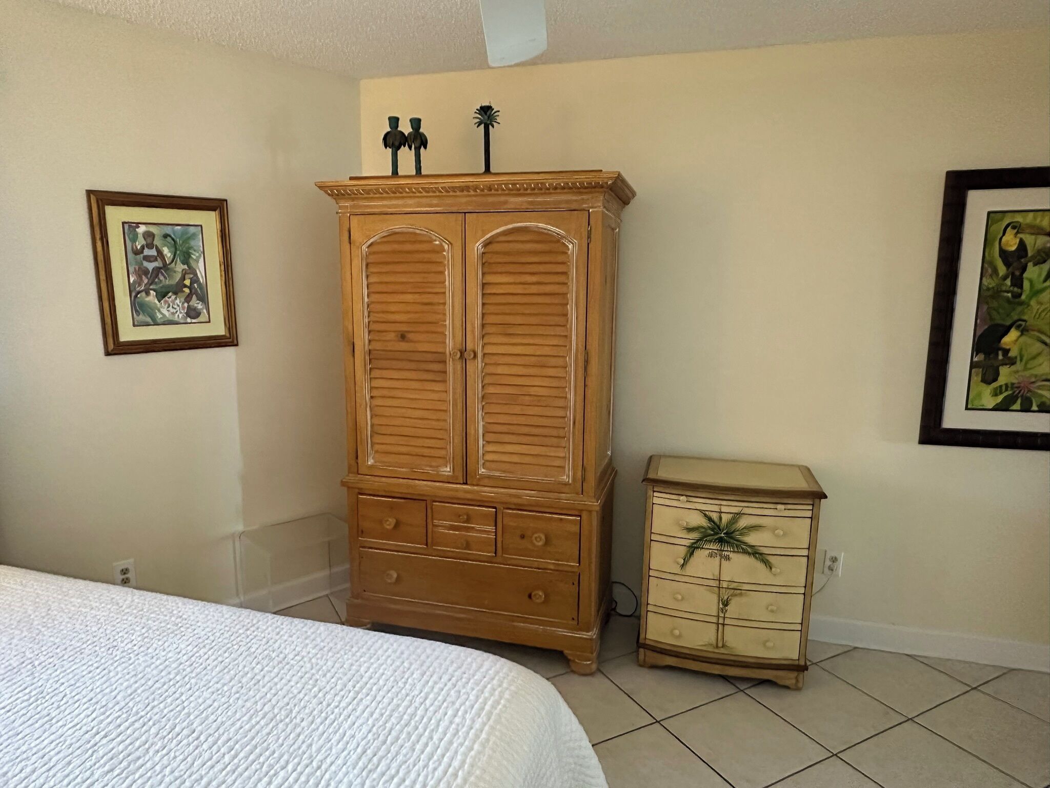 A bedroom with a bed, a wooden wardrobe, a small chest of drawers, and two framed pictures on the wall.