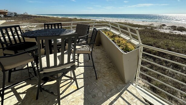A balcony with a table and chairs, overlooking a beach with a fence and grassy area. - Gulf Front Condo with Stunning Views! (Pensacola)