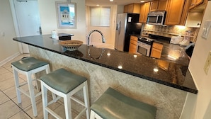 A modern kitchen with a central island, bar stools, a refrigerator, stove, microwave, and a window with blinds. - Gulf Front Condo with Stunning Views! (Pensacola)