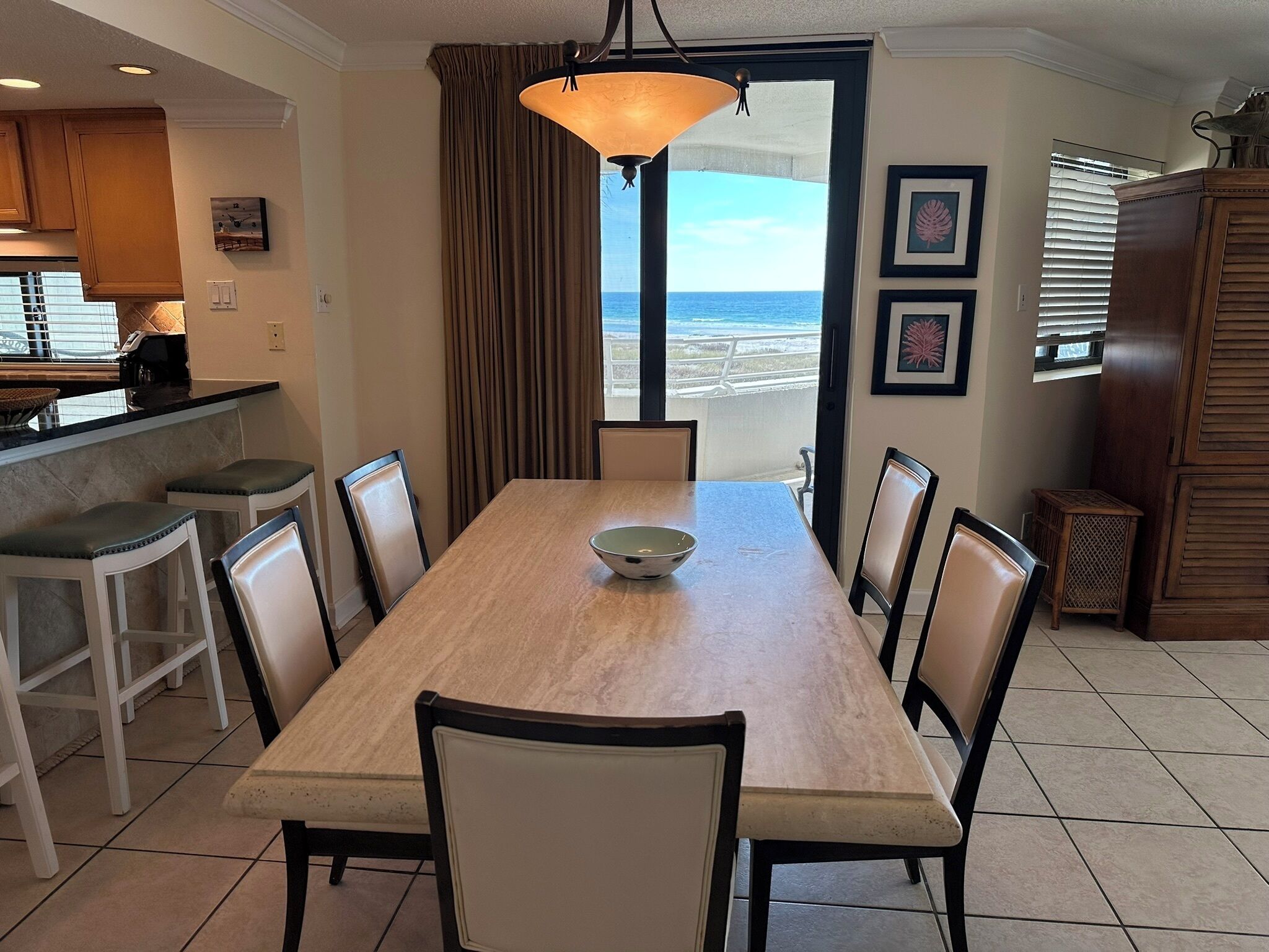 A dining area with a large table, chairs, a bowl, a pendant light, and a view of the ocean through a window.