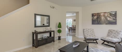 A spacious living room with a dark wooden coffee table, two patterned armchairs, a dark console table, and a large mirror. A framed painting adorns the wall, and a potted plant adds greenery.