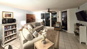 A living room with a sofa, a coffee table with a plant, a bookshelf, a TV, and a ceiling fan.
