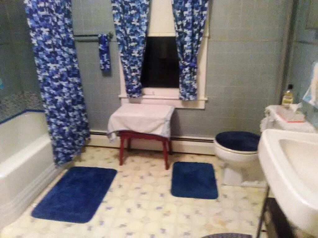 A small bathroom with a bathtub, toilet, and sink. There are blue mats on the floor and a window with curtains.