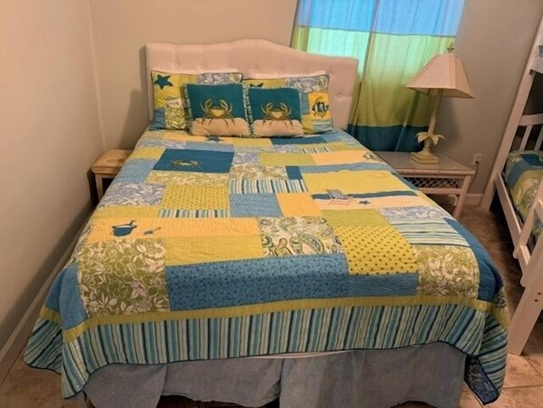 Un lit recouvert d’une courtepointe aux motifs et aux couleurs variés, une table de chevet avec une lampe, et une fenêtre avec des rideaux.