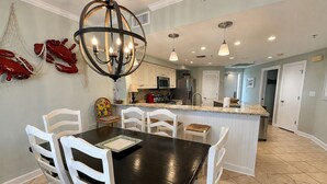A modern kitchen with white cabinetry, a central island, and a dining area with white chairs. - Gulf Front Condo with Stunning Views! (Pensacola)