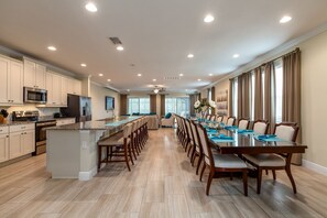 A spacious kitchen with a central island, a dining area with a long table and chairs, and a large window providing natural light.