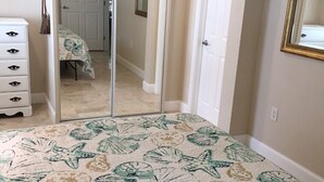 Un dormitorio con un espejo grande, una cama con colcha estampada de flores, un aparador y una puerta que da a otra habitación.