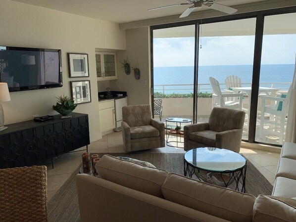 A living room with a sofa, armchairs, a coffee table, a flat-screen TV, and a balcony with a view of the ocean.