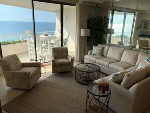 Un salón amplio con un sofá color beige, dos sillones y una mesa de centro de vidrio. Un ventanal ofrece vistas al océano.