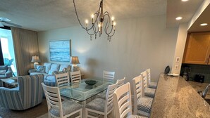 A modern dining area with a glass table, white chairs with patterned cushions, a chandelier, and a view of the ocean through a large window. - Gulf Front Condo with Stunning Views! (Pensacola)