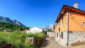 Une rue étroite bordée de bâtiments traditionnels en pierre, dont l’un est coiffé d’un toit de tuiles rouges et possède une base en pierre, et qui mène vers une chaîne de montagnes.