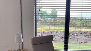 Une pièce avec un plancher en bois, un fauteuil gris recouvert de tissu et orné d’un coussin à motif de cerf, une petite table ronde avec une lampe, et une fenêtre avec des stores donnant sur un jardin.