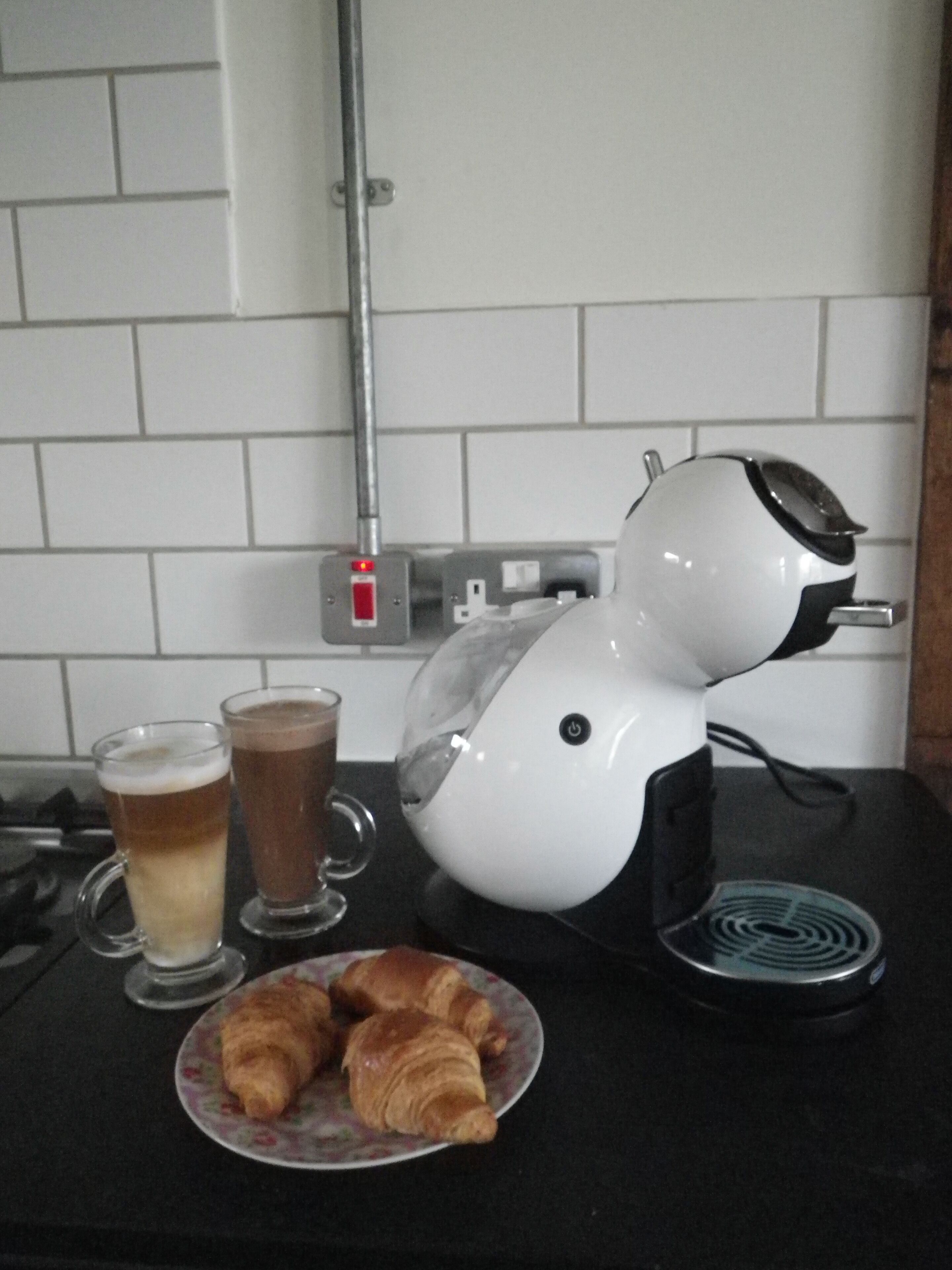 A kitchen counter with a coffee maker, two glasses of coffee, and a plate of croissants.