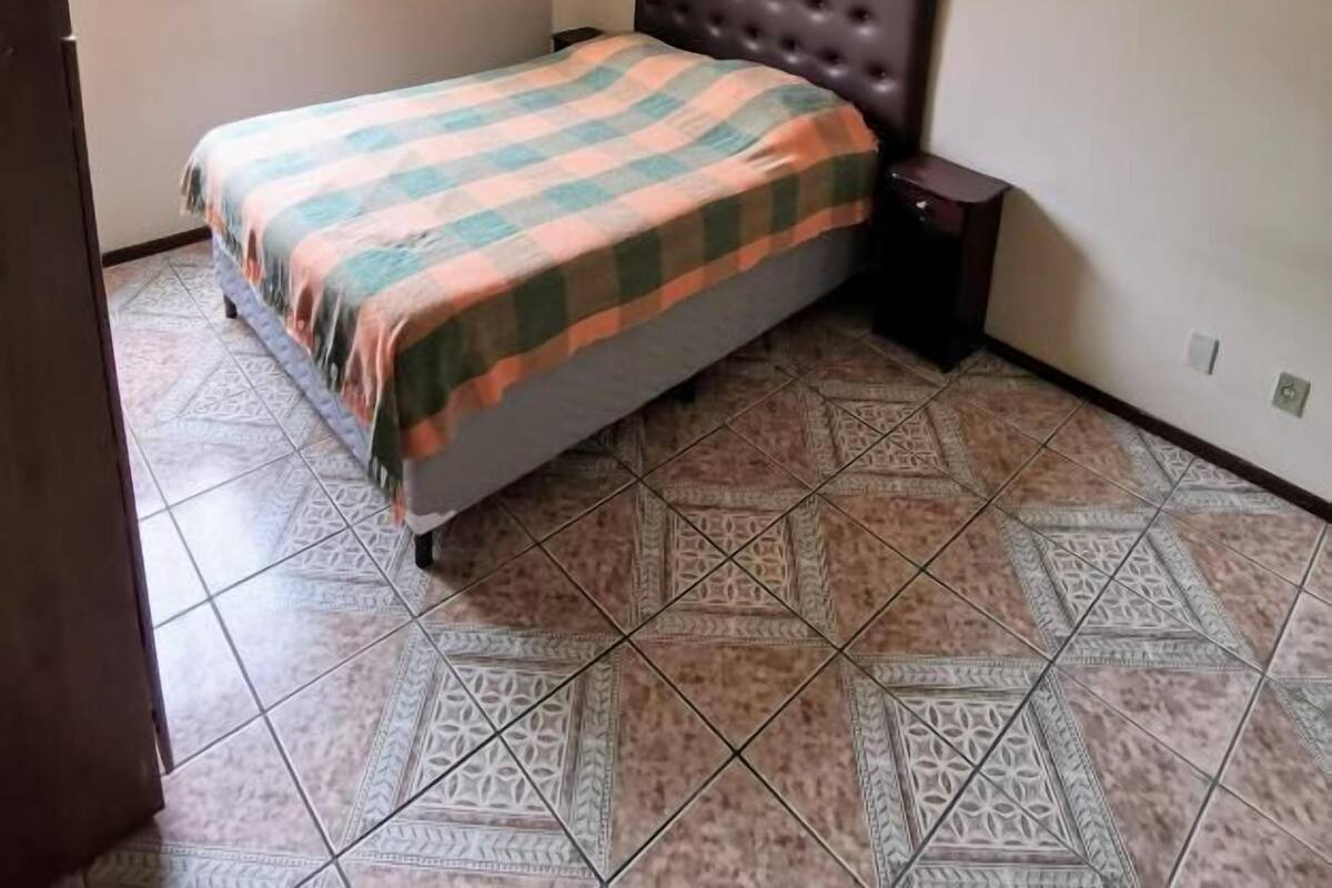 A bedroom with a bed, a ceiling fan, a window with curtains, and patterned floor tiles.