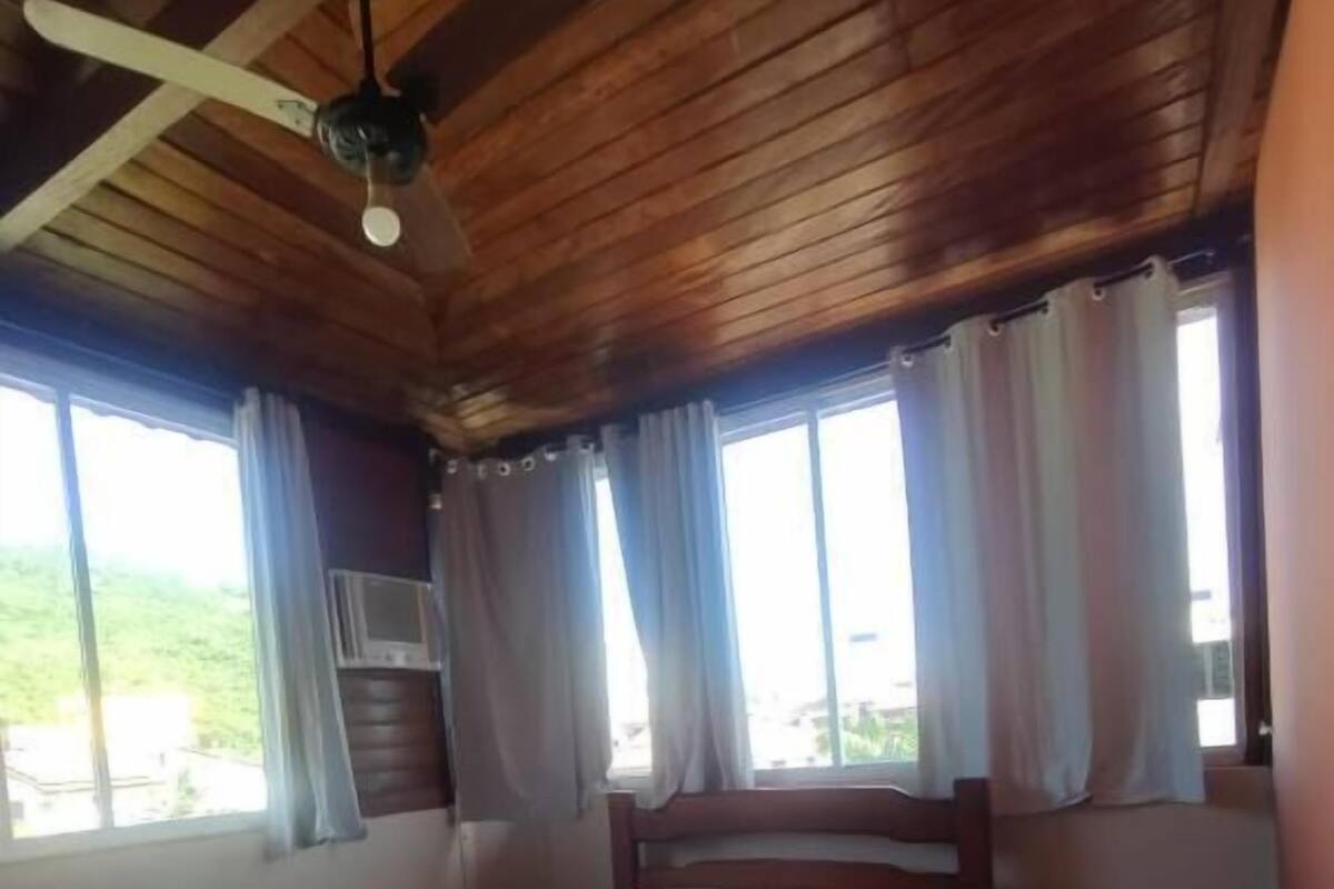 A bedroom with a wooden ceiling fan, a bed with a headboard, and large windows with curtains.