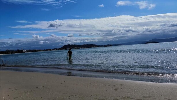 A beach with a person standing in the water, a clear sky, and distant mountains. - Apart da Cachoeira (Florianópolis)