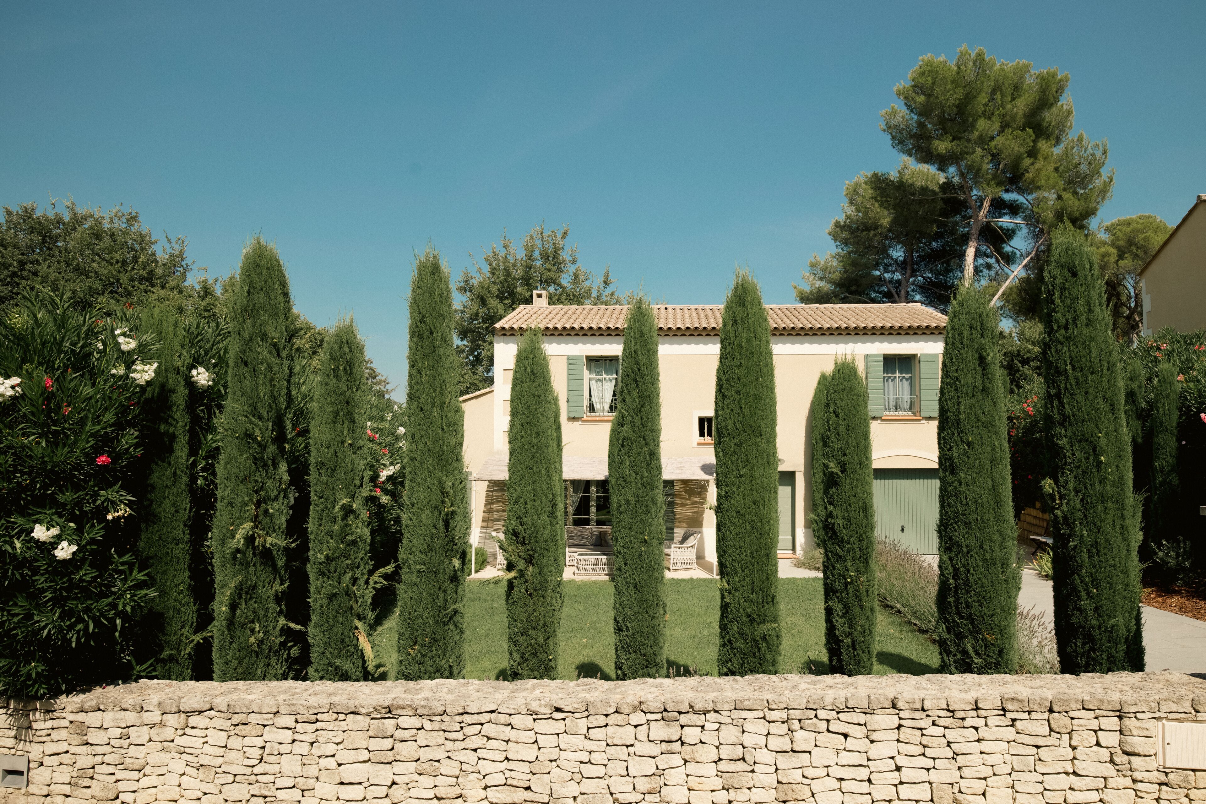 A two-story house with a stone wall in the foreground, surrounded by tall, narrow trees.