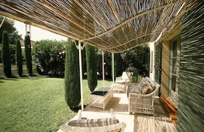 Un patio couvert aménagé avec des meubles en osier, une table en verre et une vue sur une pelouse et un jardin bien entretenus.
