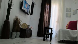 A living room with a bed, a chair, a painting, and a window with curtains.