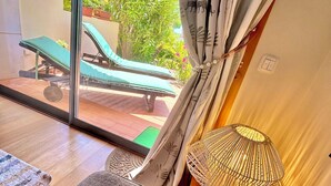 Habitación con vista a un jardín, una silla de descanso, una lámpara estampada y suelo de madera.