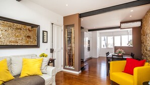 Un saló modern amb un sofà blanc, coixins grocs i un butacó groc. Hi ha un quadre abstracte gran a la paret, una porta de vidre que condueix a una altra habitació i una zona de menjador amb taula i cadires.