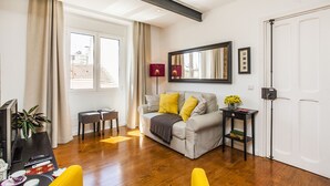 Un saló modern amb un sofà, una taula de centre i una televisió. Hi ha una finestra amb cortines, una porta i una taula petita amb un vas de flors.