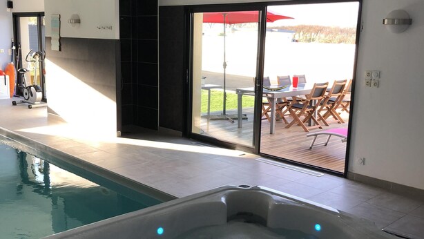 A modern indoor swimming pool with a hot tub, a patio area with outdoor furniture, and a view of the outdoors.