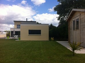 A modern two-story house with a flat roof, a large window on the second floor, and a small garden area with a lawn and a few plants.